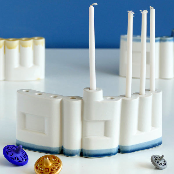 **Innovative “Shalom” Hanukkah Menorah (Chanukiah)**
A unique arc-shaped menorah spelling the Hebrew word *Shalom* (שלום), slip-cast by us in our Tel Aviv studio. Off-white clay, matte off-white glaze, with a subtle blue accent along the base—blending tradition with a fresh, modern look.