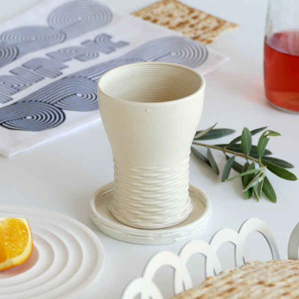 This elegant Kiddush cups set has clean shape with weaving pattern at its bottom - created in a unique method by a clay 3D Printer. The set includes one large Kiddish wine goblet with a plate, and six matching small cups.