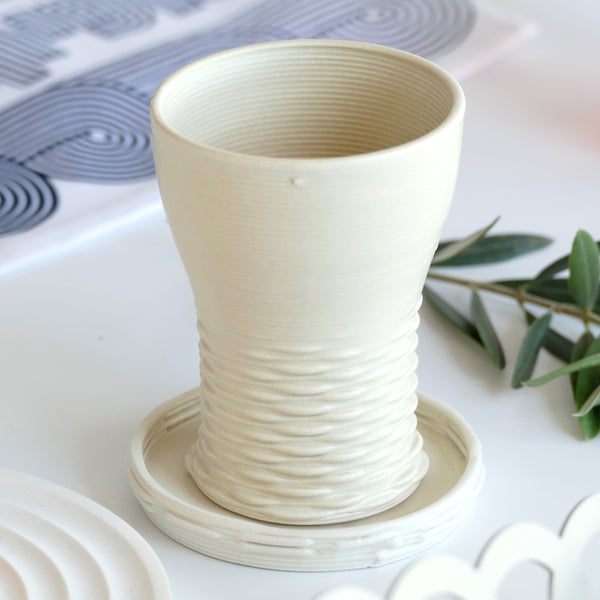 This elegant Kiddush cups set has clean shape with weaving pattern at its bottom - created in a unique method by a clay 3D Printer. The set includes one large Kiddish wine goblet with a plate, and six matching small cups.